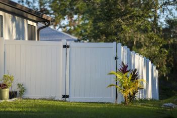White vinyl backyard fence and gate beside a lawn with potted plants and tall trees in the background
